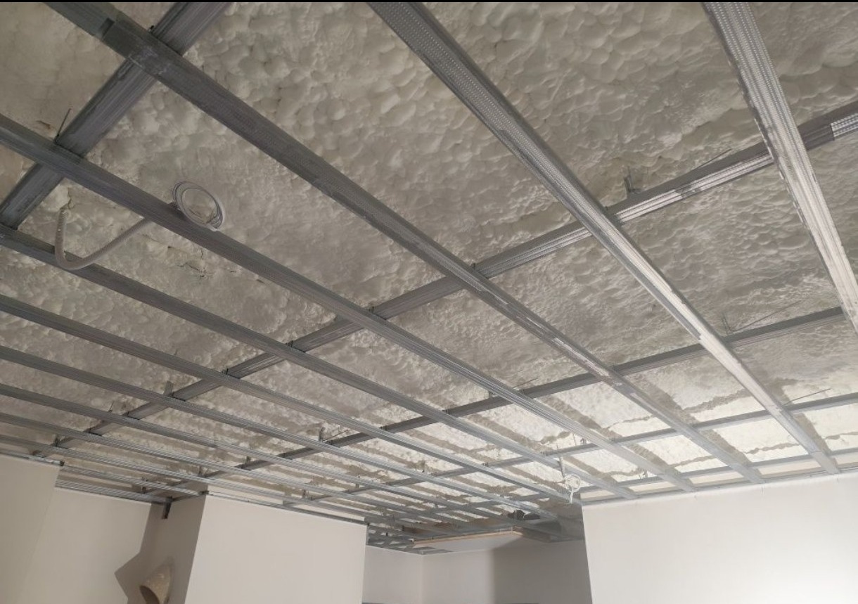 Sufit podwieszany z metalową konstrukcją i pianką izolacyjną, widok z dołu. Widoczne elementy instalacji elektrycznej. Jasne wnętrze w trakcie prac wykończeniowych.
