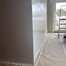 SC CONSTRUCK SPÓŁKA Z OGRANICZONĄ ODPOWIEDZIALNOŚCIĄ - Ściana po szpachlowaniu, częściowo pomalowana na biało, z widocznymi śladami szpachli. Podłoga zabezpieczona folią malarską. W tle okno z drabiną.
