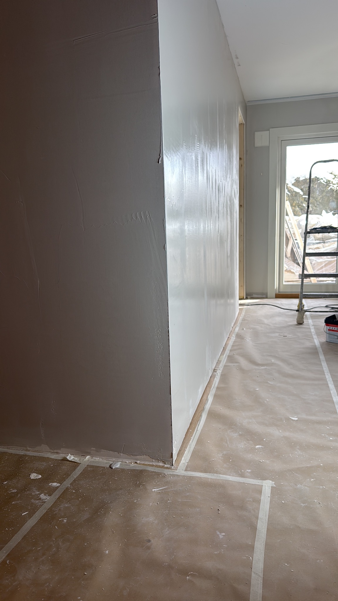 Ściana po szpachlowaniu, częściowo pomalowana na biało, z widocznymi śladami szpachli. Podłoga zabezpieczona folią malarską. W tle okno z drabiną.