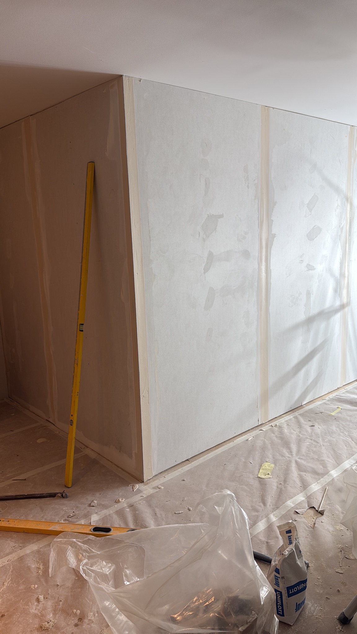 Ściana z płyt karton-gips, częściowo wyszpachlowana, z widocznymi narzędziami (poziomica) i workiem gipsu na podłodze zabezpieczonej folią malarską.