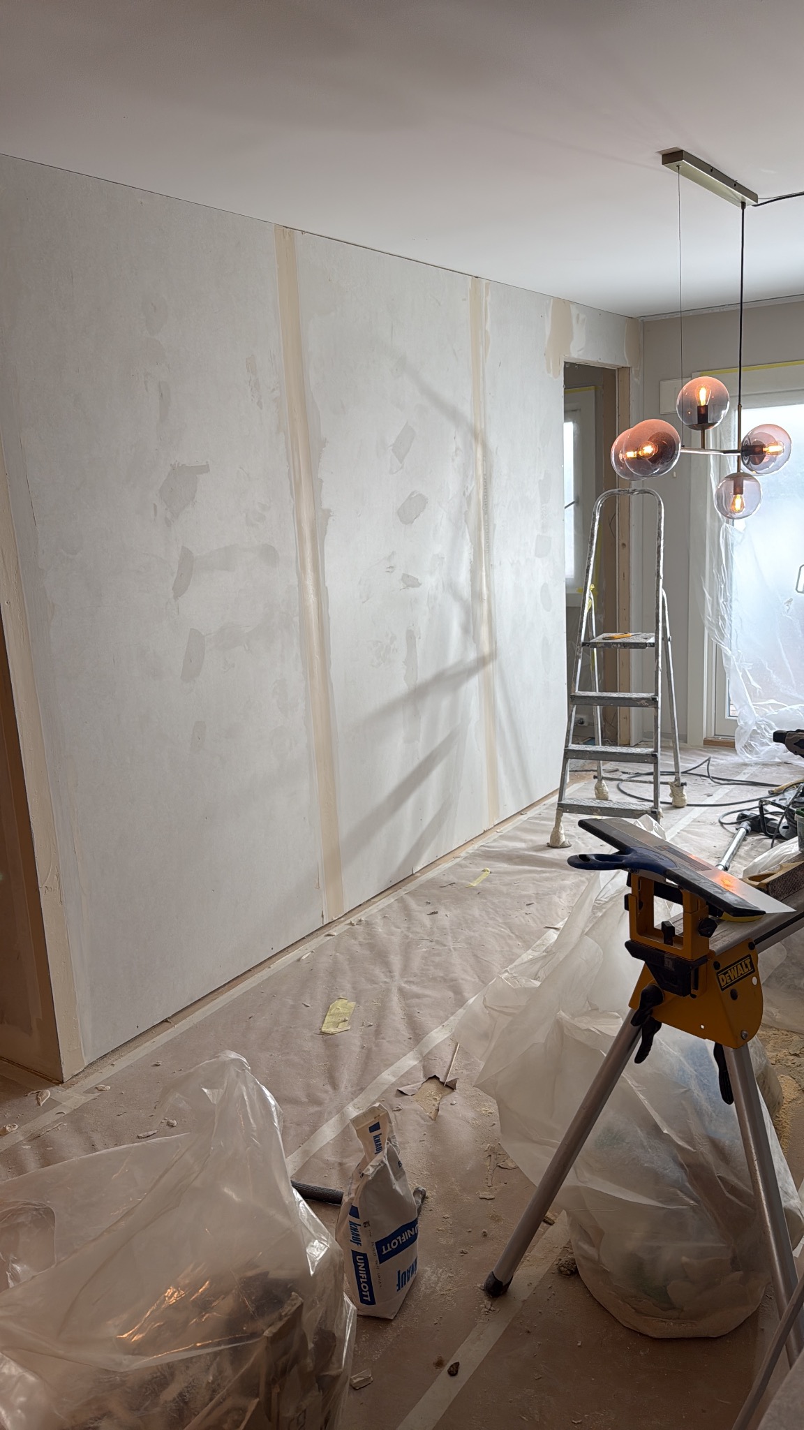 Ściana po szpachlowaniu w Rogoźnie, przygotowana do malowania. Widoczne taśmy maskujące, drabina i narzędzia. Nowoczesna lampa sufitowa.