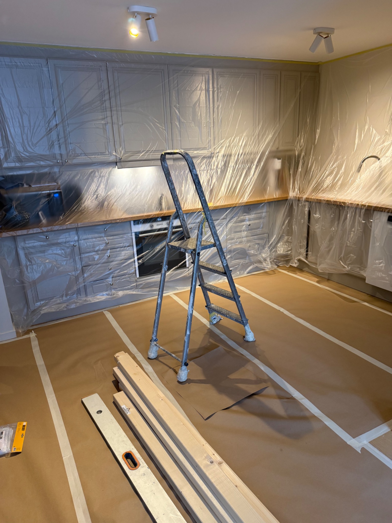 Przygotowana kuchnia do malowania: zabezpieczone folią meble i podłoga, drabina, poziomica i deski na podłodze. Widok na fronty szafek i sprzęty AGD.