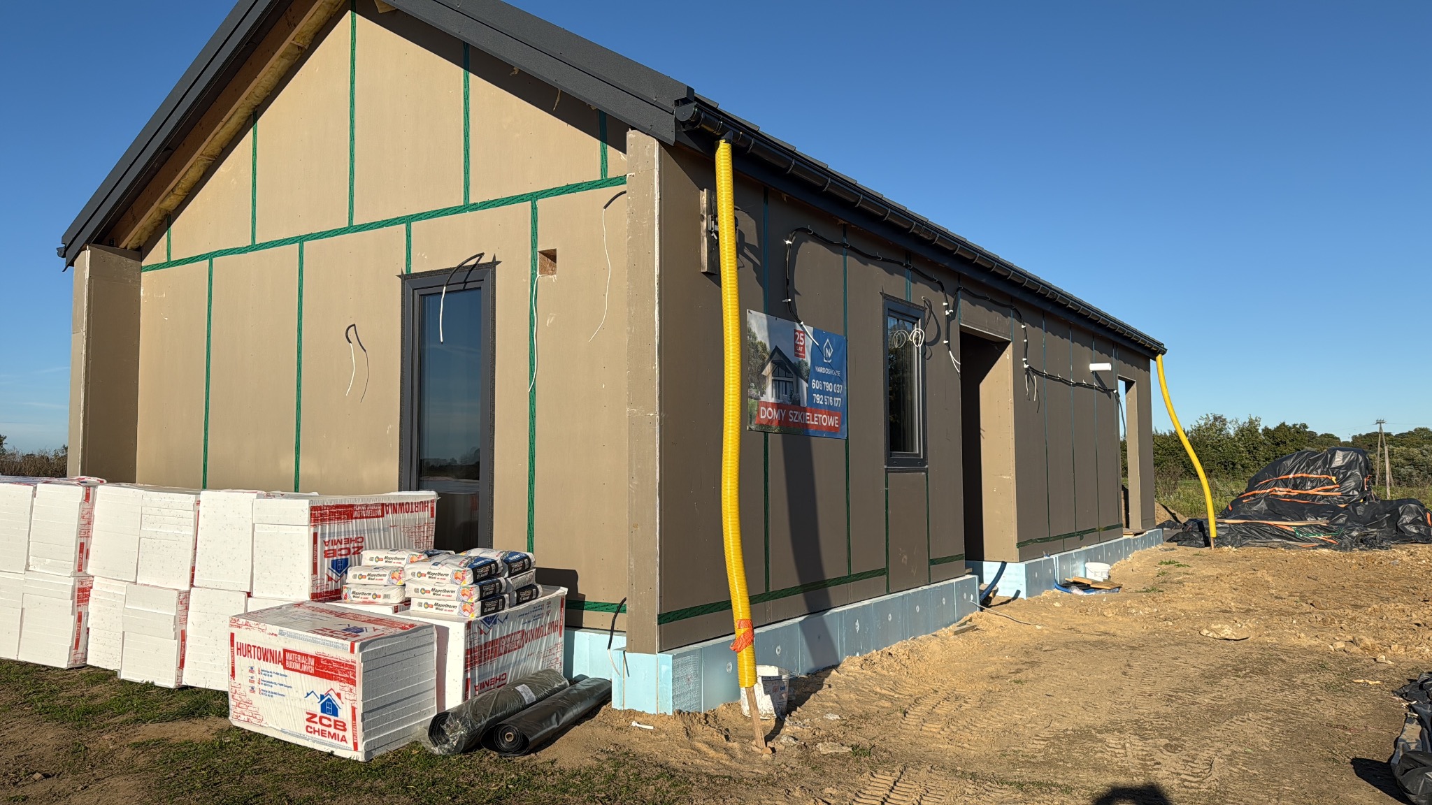 Surowy dom szkieletowy w trakcie budowy, z widocznymi materiałami izolacyjnymi i elementami konstrukcyjnymi. Żółty przewód wentylacyjny przy ścianie. Błękitne niebo w tle.
