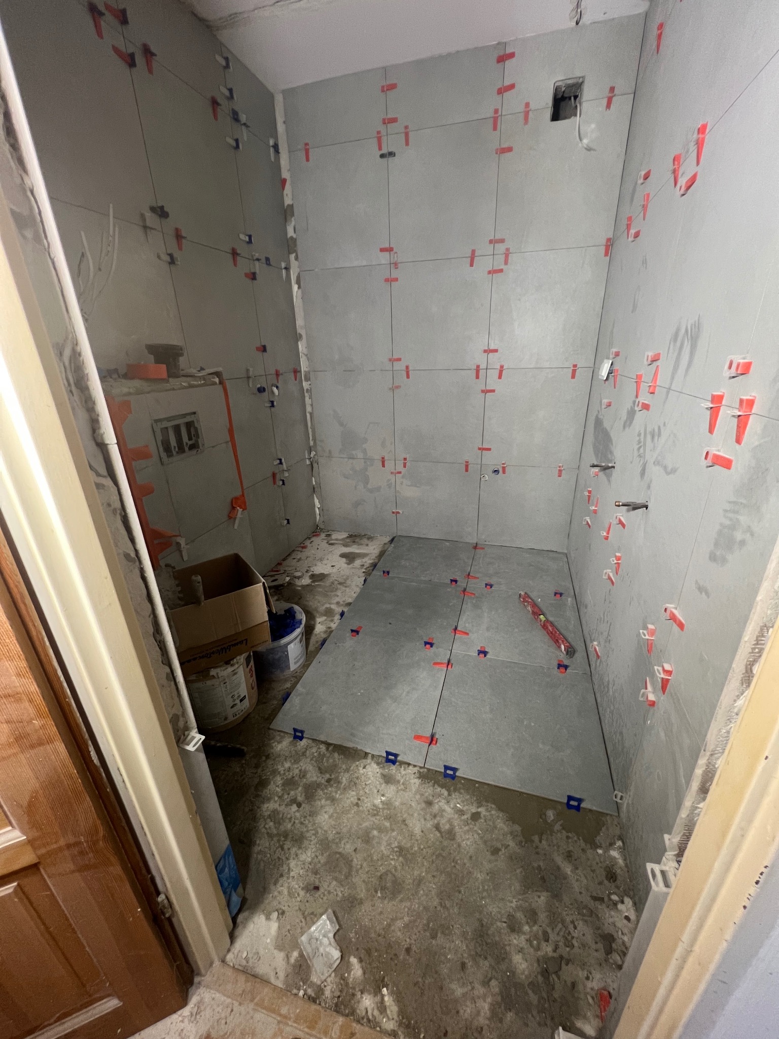 Układanie szarych płytek w łazience z użyciem poziomicy i klipsów dystansowych. Widoczne niedokończone ściany i podłoga. W tle farby i karton.