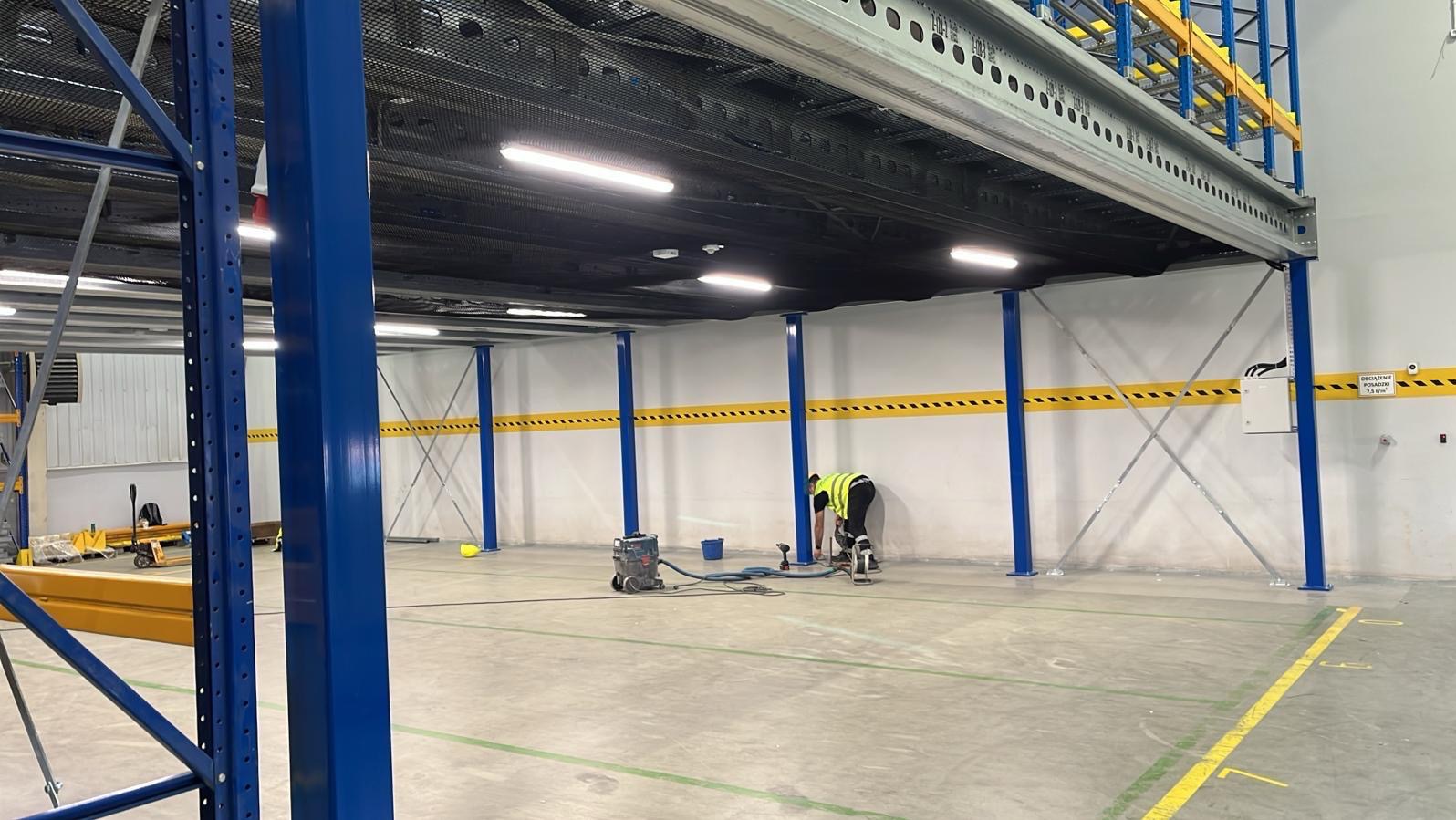 Montaż antresoli magazynowej: pracownik w kamizelce, szlifuje posadzkę pod konstrukcję. Widoczne niebieskie słupy, żółte pasy na ścianach i oświetlenie LED.