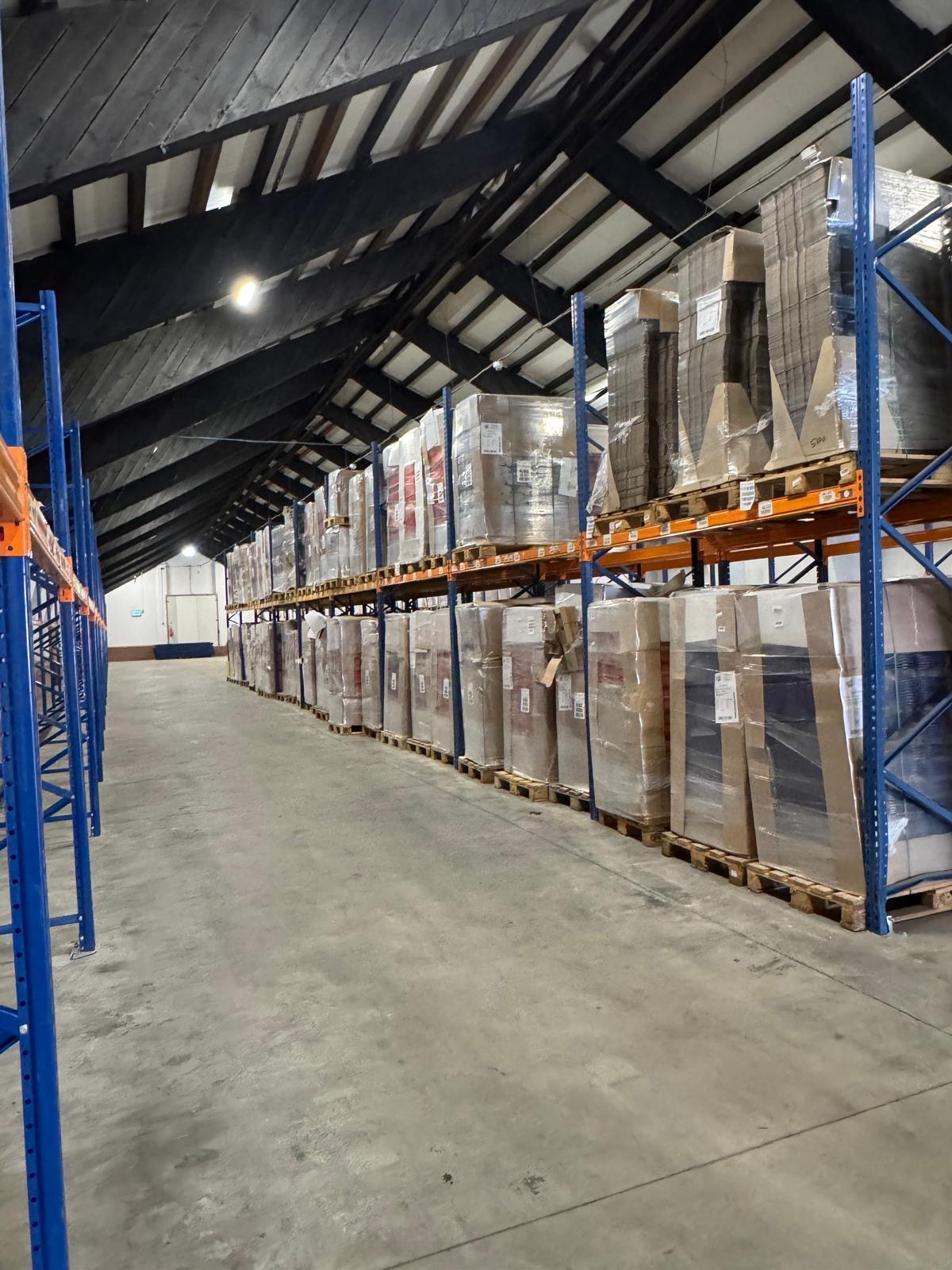 Magazyn z wysokimi regałami, na których składowane są zapakowane towary. Widoczne niebieskie ramy regałów i pomarańczowe poprzeczki. Hala magazynowa z ciemnym stropem.