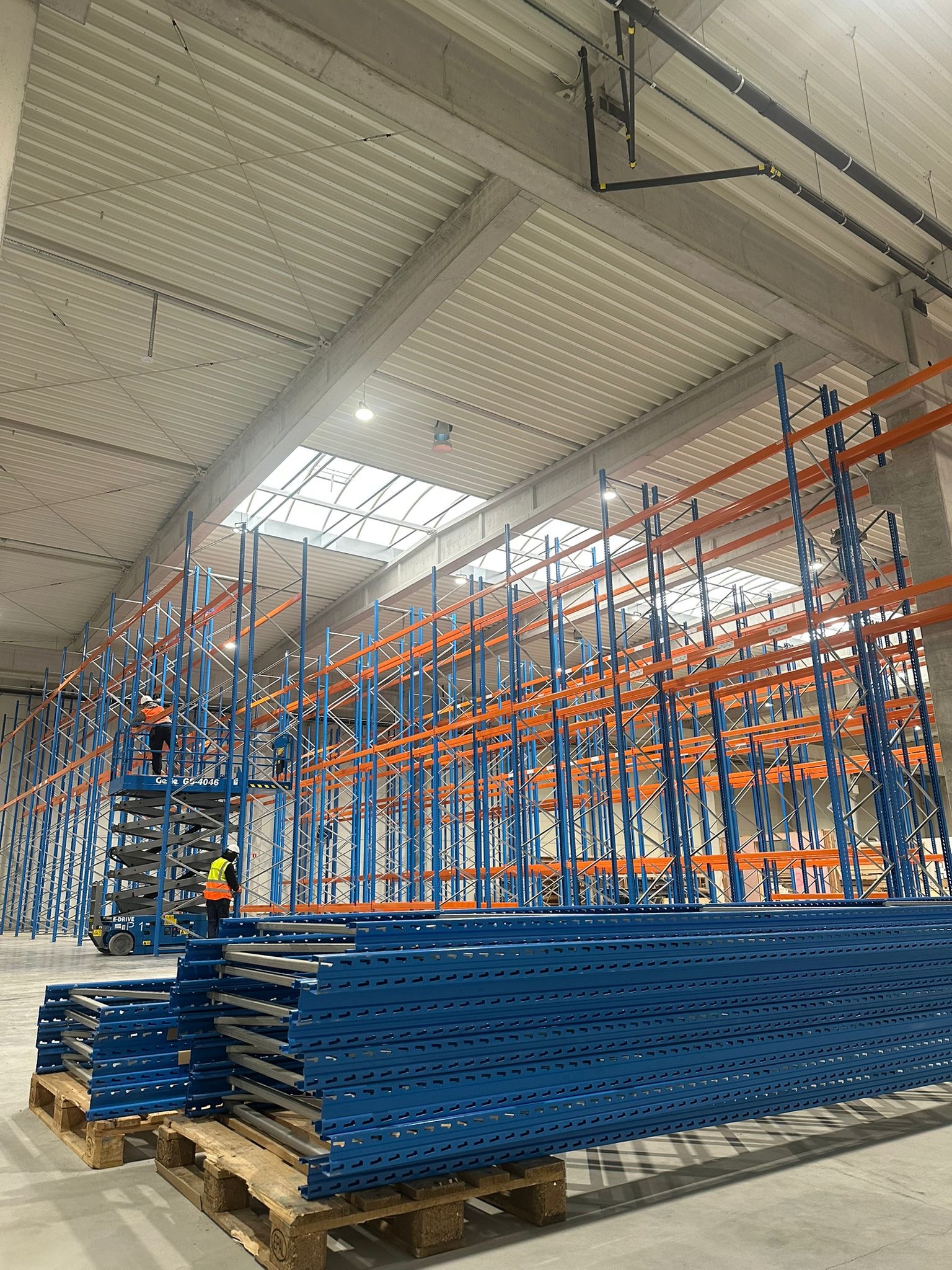 Montaż regałów magazynowych w hali: pracownicy na podnośniku instalują niebiesko-pomarańczowe elementy. Na pierwszym planie stos niebieskich belek na paletach.