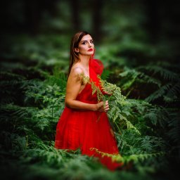 Ewa Gorzeń - Kobieta w czerwonej sukni, otoczona paprociami w lesie, trzymająca gałązkę paproci. Ujęcie z dołu, z ciemnym tłem i mocnym kontrastem kolorów.