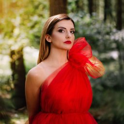 Ewa Gorzeń - Portret kobiety w czerwonej, tiulowej sukni na tle leśnej scenerii. Uwagę przykuwa intensywna czerwień sukni i wyrazisty makijaż modelki, kontrastujące z zielenią tła.
