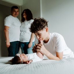 Ewa Gorzeń - Rodzinne ujęcie: nastolatek całuje stópkę niemowlaka leżącego na łóżku, w tle rodzice w białych koszulkach. Jasne, naturalne światło, minimalistyczne wnętrze.