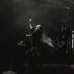 Ewa Gorzeń - Czarno-białe zdjęcie wokalistki z uniesioną ręką na scenie podczas koncertu, w tle gitarzysta i światła sceniczne. Dynamiczne ujęcie z występu na żywo.