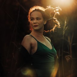 Ewa Gorzeń - Portret kobiety w zielonej sukience, oświetlonej ciepłym światłem zachodzącego słońca, z włosami upiętymi w kok, na tle wysokich traw i zarośli.