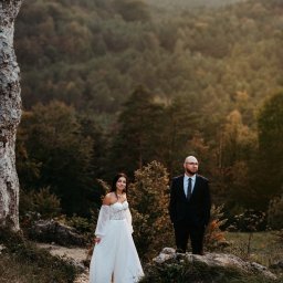 Ewa Gorzeń - Para młoda w plenerze: panna młoda w białej sukni z odkrytymi ramionami i pan młody w czarnym garniturze na tle leśnego krajobrazu i skał. Romantyczna sceneria.