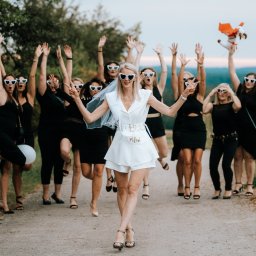 Ewa Gorzeń - Uśmiechnięta panna młoda w bieli, otoczona druhnami w czerni i okularach w kształcie serc, świętuje wieczór panieński na leśnej drodze.