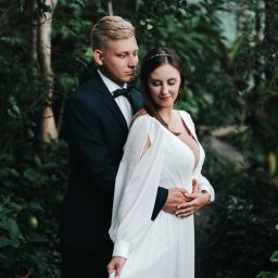 Ewa Gorzeń - Para młoda w objęciach, on w granatowym smokingu z muchą, ona w białej sukni z długim rękawem, na tle zieleni. Romantyczna sesja w plenerze.