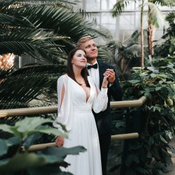 Ewa Gorzeń - Para w strojach ślubnych, trzymająca się za ręce w tropikalnej scenerii palmiarni w Lędzinach. Panna młoda w białej sukni, pan młody w smokingu.