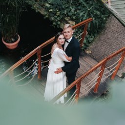 Ewa Gorzeń - Para młoda na drewnianym mostku w otoczeniu zieleni. Panna młoda w białej sukni, pan młody w czarnym garniturze. Ujęcie z góry, z delikatnym rozmytym pierwszym planem.