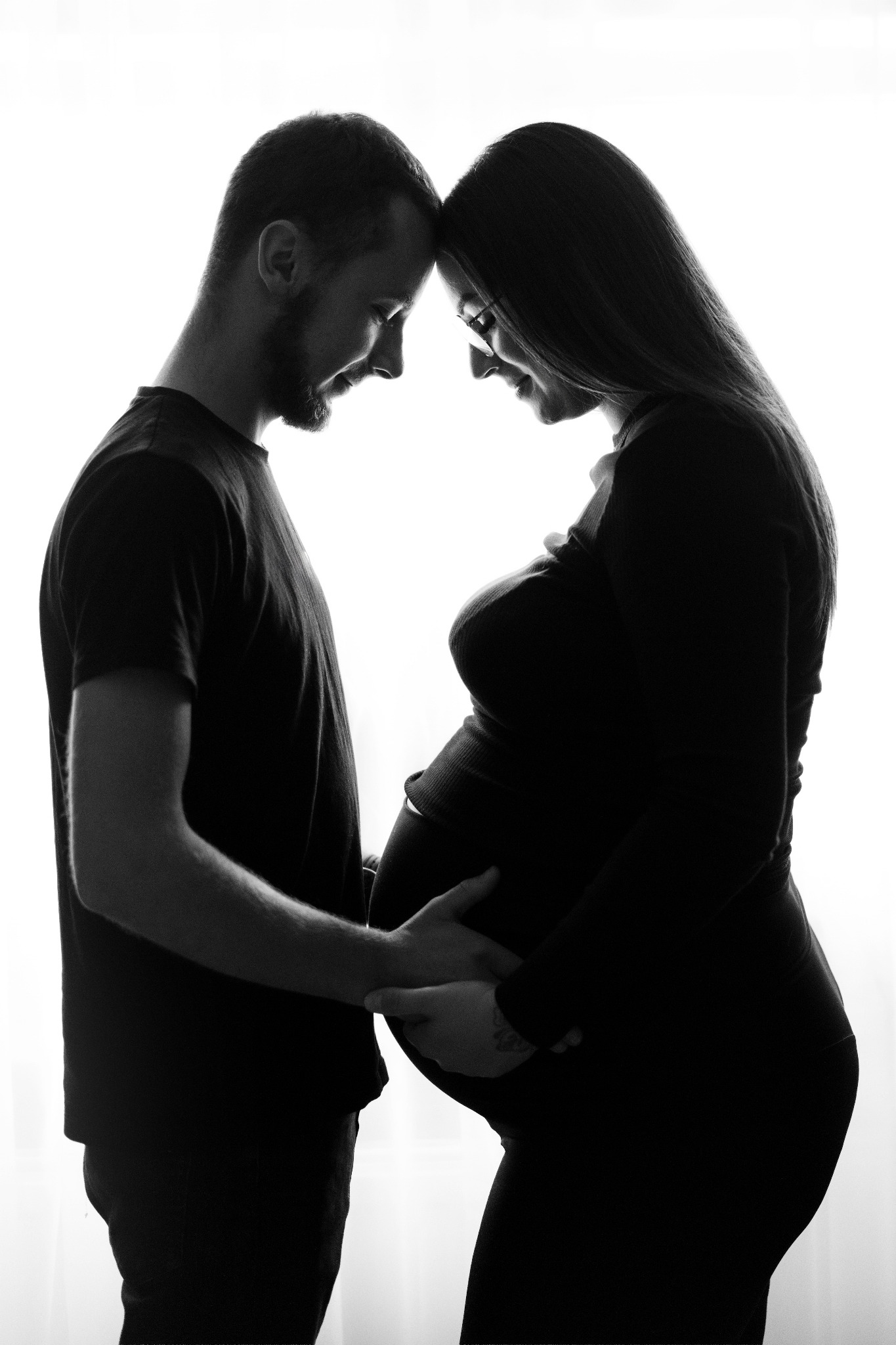 Czarno-biała fotografia sylwetek kobiety w ciąży i mężczyzny, stykających się czołami, dłonie na brzuchu. Delikatne światło podkreśla intymność i oczekiwanie.