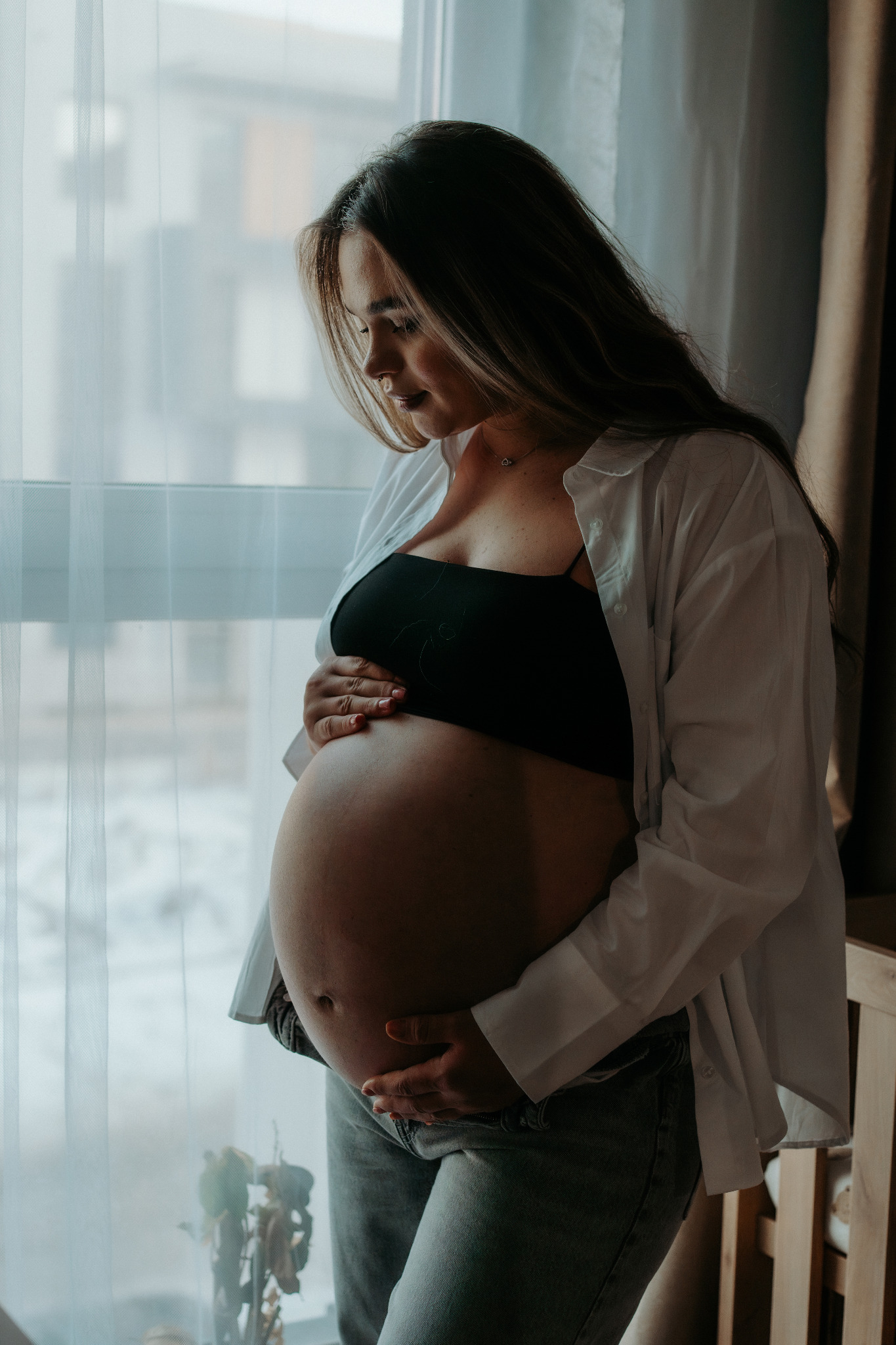 Kobieta w ciąży, w jasnej koszuli i topie, delikatnie obejmuje brzuch na tle okna z firanką. Subtelne światło podkreśla naturalność i intymność momentu.