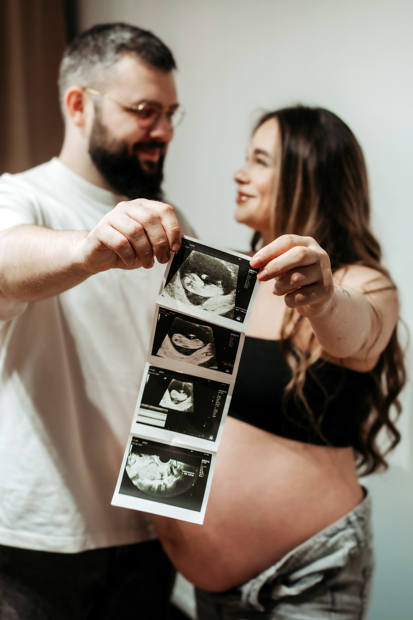 Uśmiechnięta para trzymająca wydruk USG, kobieta w ciąży w czarnym topie i dżinsach, mężczyzna w białej koszulce, ciepłe oświetlenie, neutralne tło.
