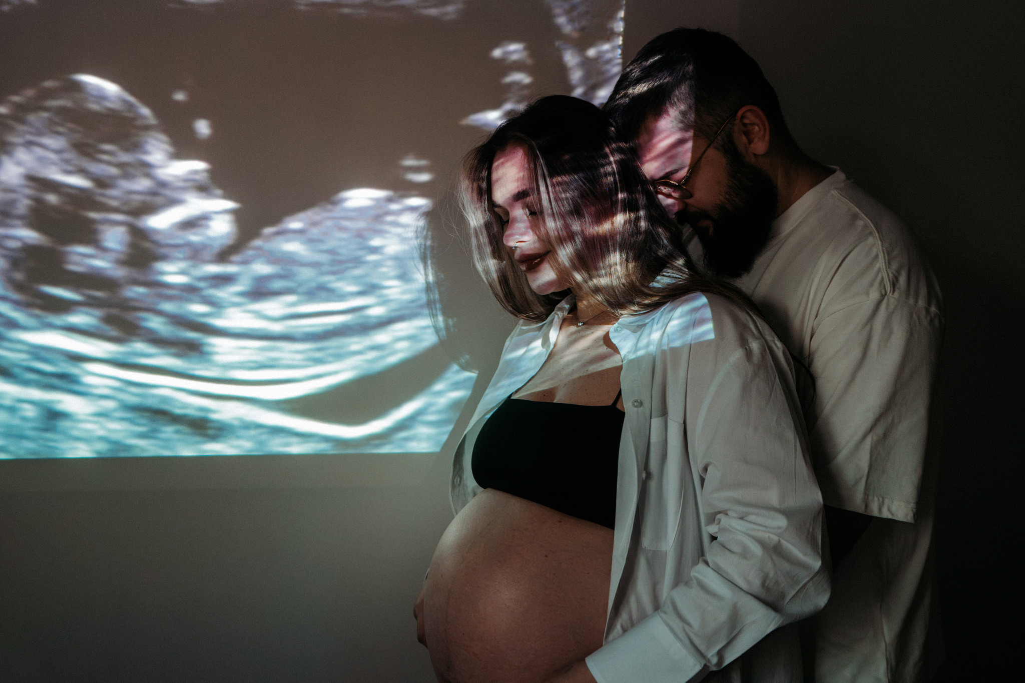 Czułe ujęcie przyszłych rodziców w intymnej sesji ciążowej, z projekcją USG na ścianie, tworząc unikalny efekt wizualny i podkreślając bliskość oczekiwania na dziecko.