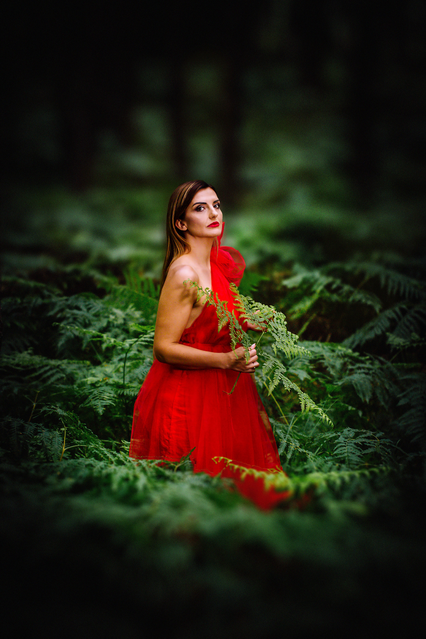 Kobieta w czerwonej sukni, otoczona paprociami w lesie, trzymająca gałązkę paproci. Ujęcie z dołu, z ciemnym tłem i mocnym kontrastem kolorów.