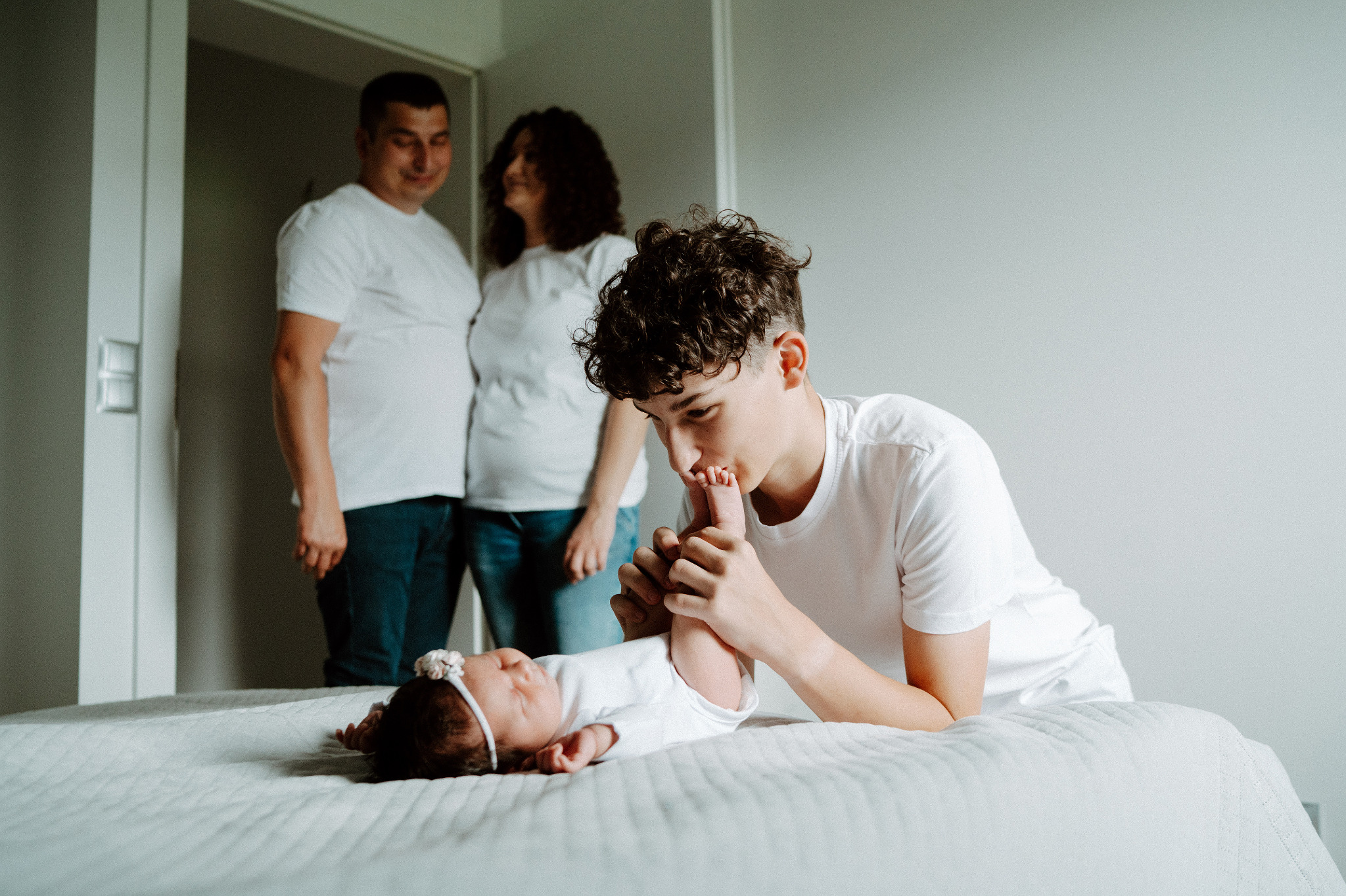 Rodzinne ujęcie: nastolatek całuje stópkę niemowlaka leżącego na łóżku, w tle rodzice w białych koszulkach. Jasne, naturalne światło, minimalistyczne wnętrze.