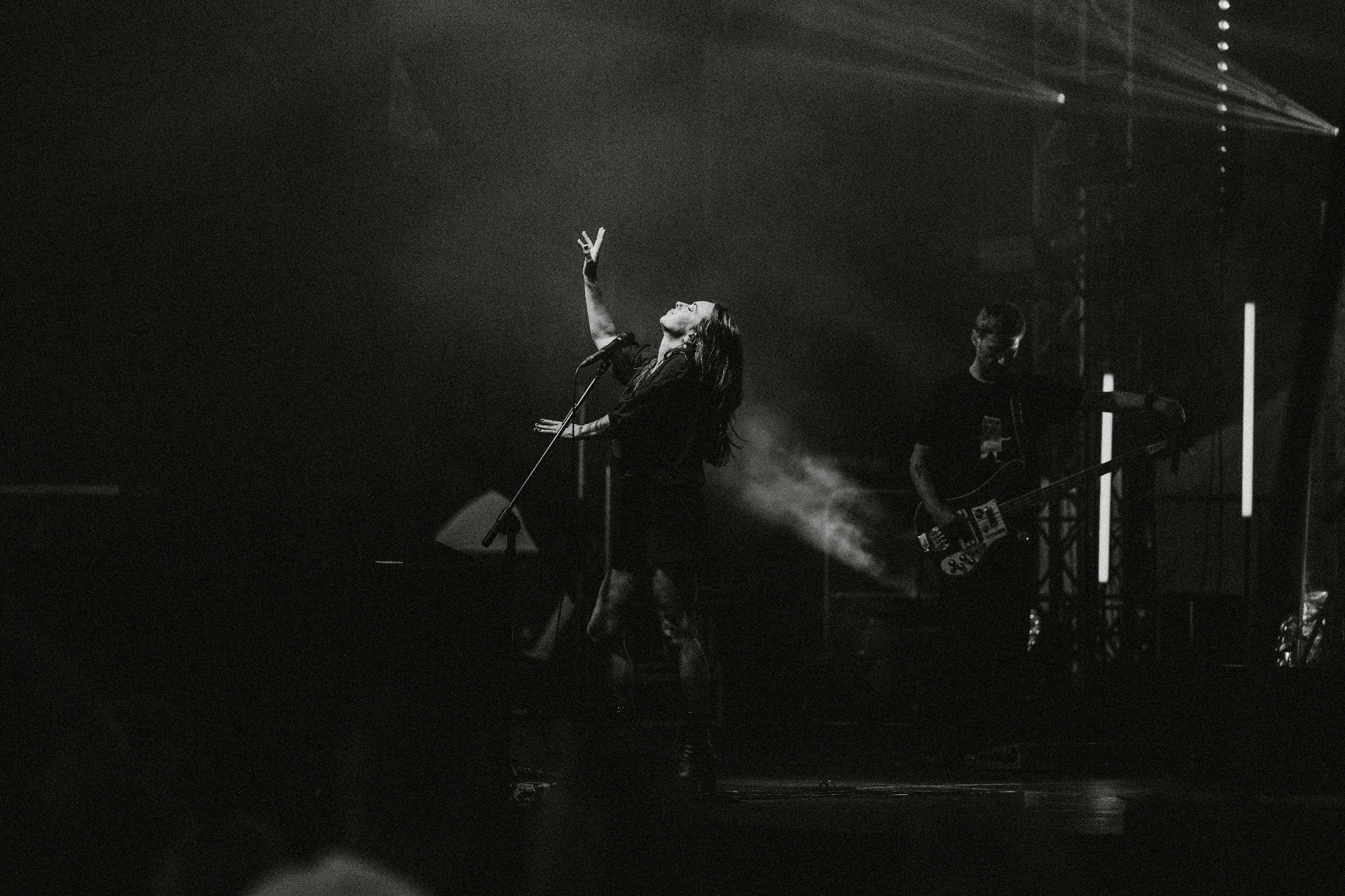 Czarno-białe zdjęcie wokalistki z uniesioną ręką na scenie podczas koncertu, w tle gitarzysta i światła sceniczne. Dynamiczne ujęcie z występu na żywo.
