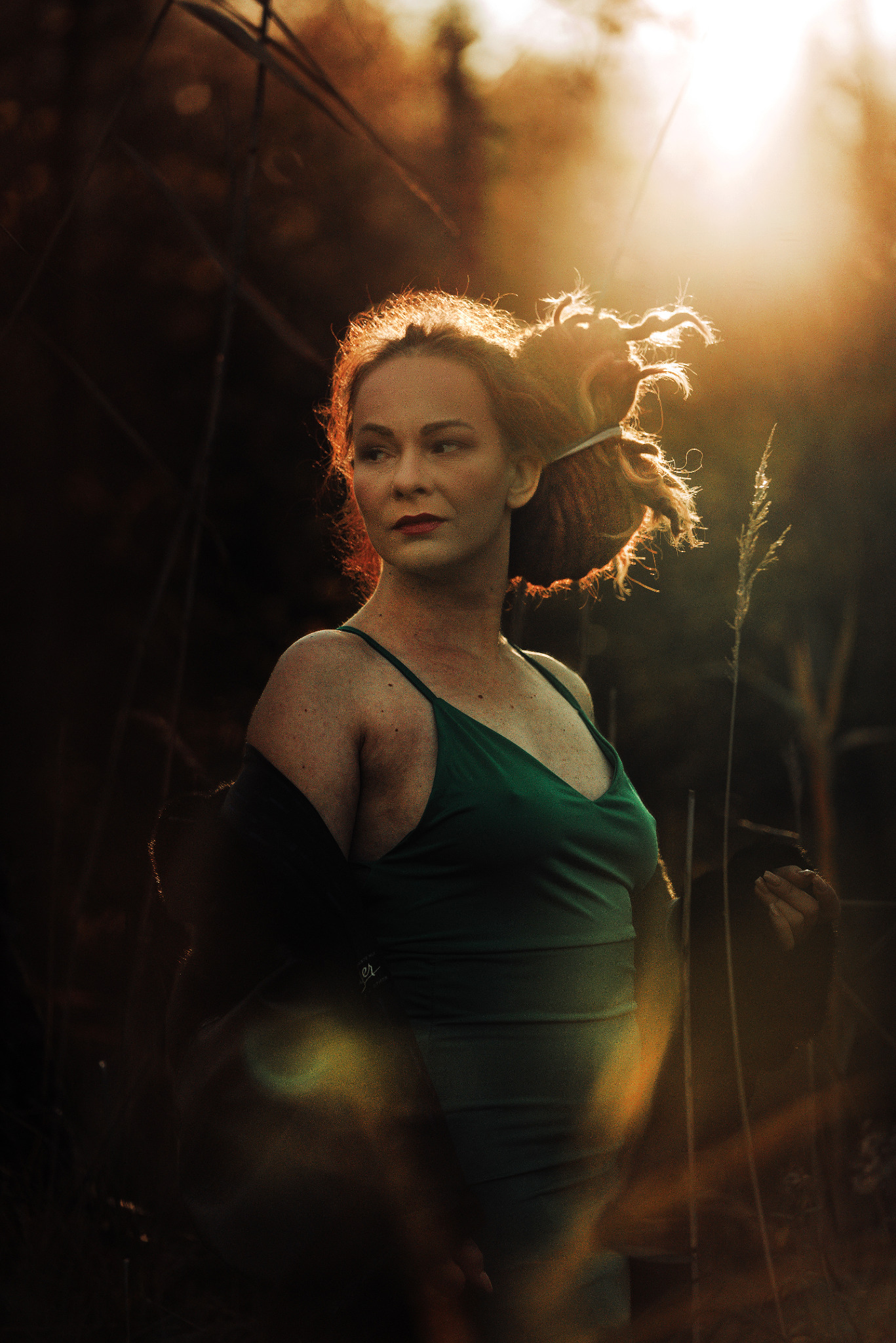Portret kobiety w zielonej sukience, oświetlonej ciepłym światłem zachodzącego słońca, z włosami upiętymi w kok, na tle wysokich traw i zarośli.