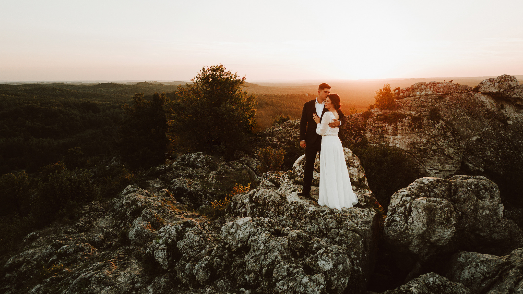 Para młoda w objęciach na skalistym wzgórzu o zachodzie słońca. Ona w białej sukni, on w czarnym garniturze. Romantyczna sceneria z widokiem na las i pomarańczowe niebo.