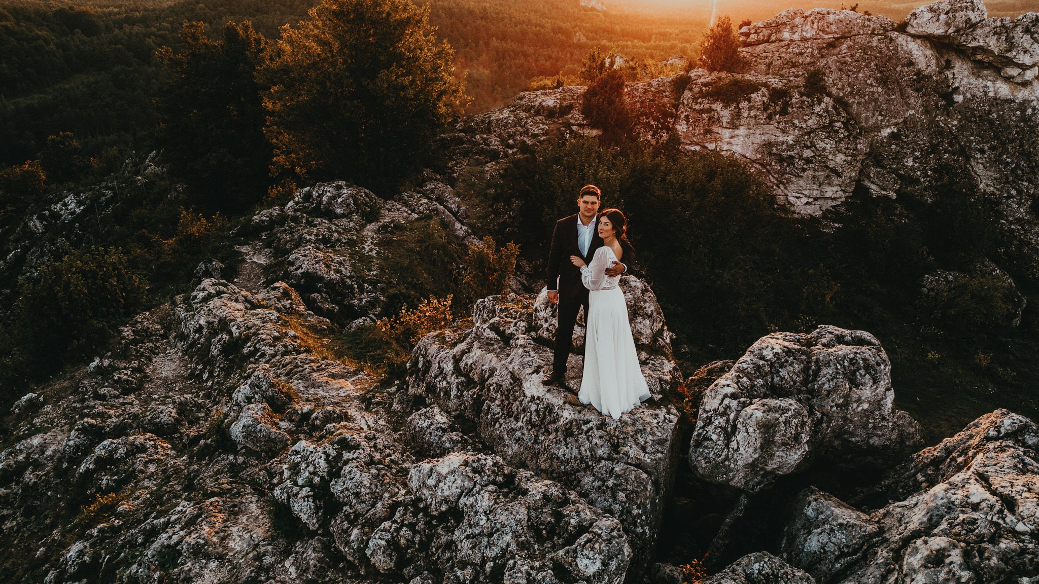 Para młoda w objęciach na skalistym wzgórzu o zachodzie słońca. Ona w białej sukni z długim rękawem, on w ciemnym garniturze. Romantyczna sceneria z widokiem na las.