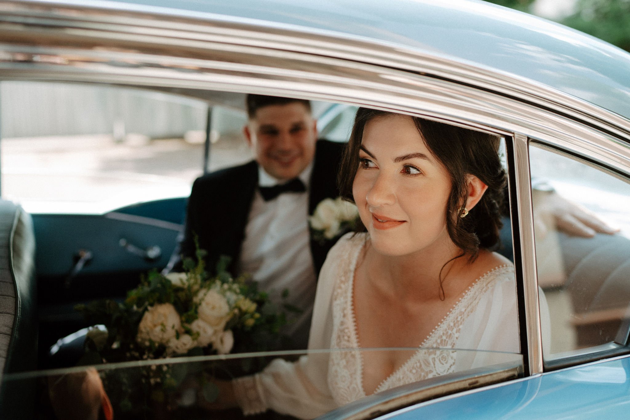 Uśmiechnięta panna młoda w sukni z koronką i bukietem w błękitnym samochodzie retro, obok pan młody w smokingu. Romantyczna sesja ślubna w stylu vintage.