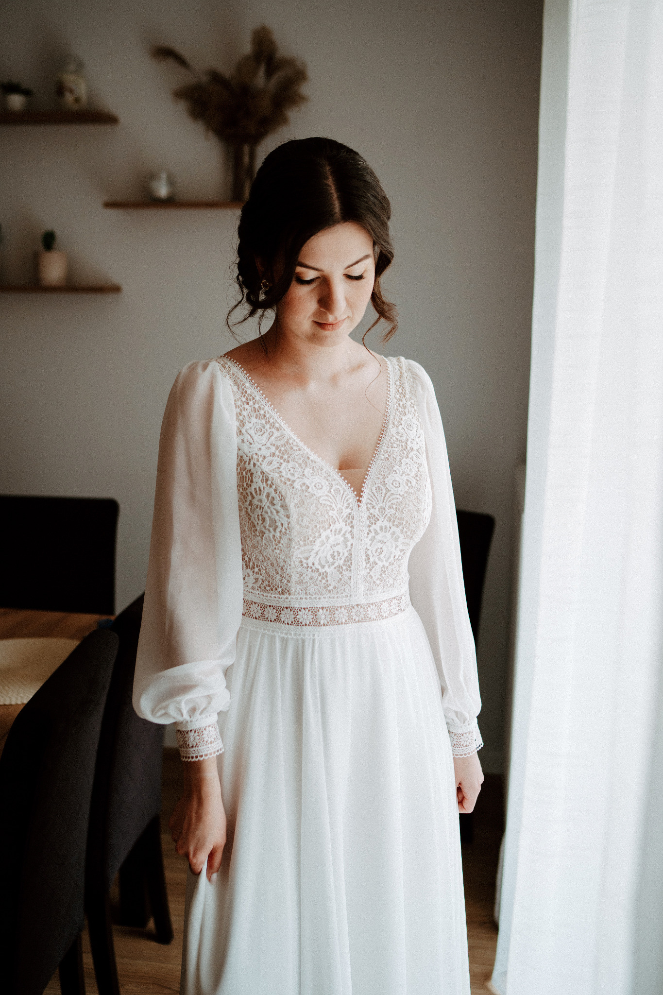 Panna młoda w koronkowej sukni ślubnej z bufiastymi rękawami, spogląda w dół, stojąc przy oknie z delikatnym światłem. W tle minimalistyczny wystrój wnętrza.