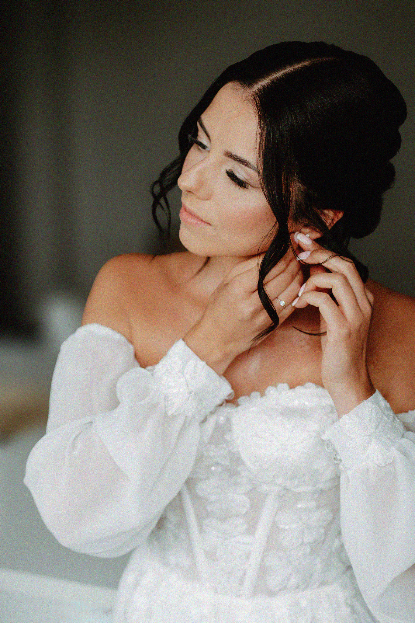 Panna młoda w białej sukni z koronkowym gorsetem, z delikatnym makijażem, zakłada kolczyk. Profil, stonowana kolorystyka, miękkie światło.