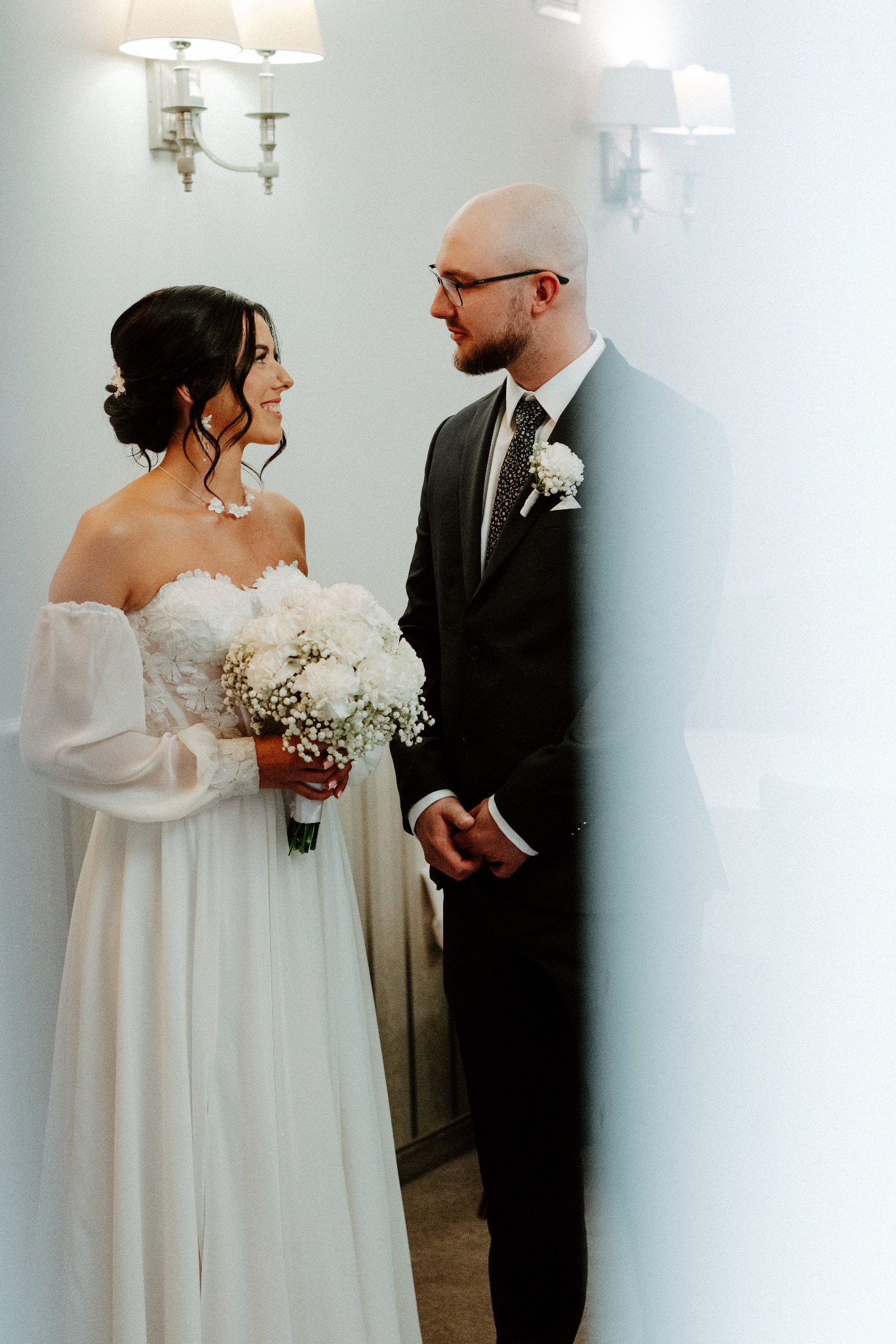 Uśmiechnięta panna młoda w białej sukni z bukietem spogląda na pana młodego w czarnym garniturze, w jasnym wnętrzu. Delikatne światło, subtelna elegancja.