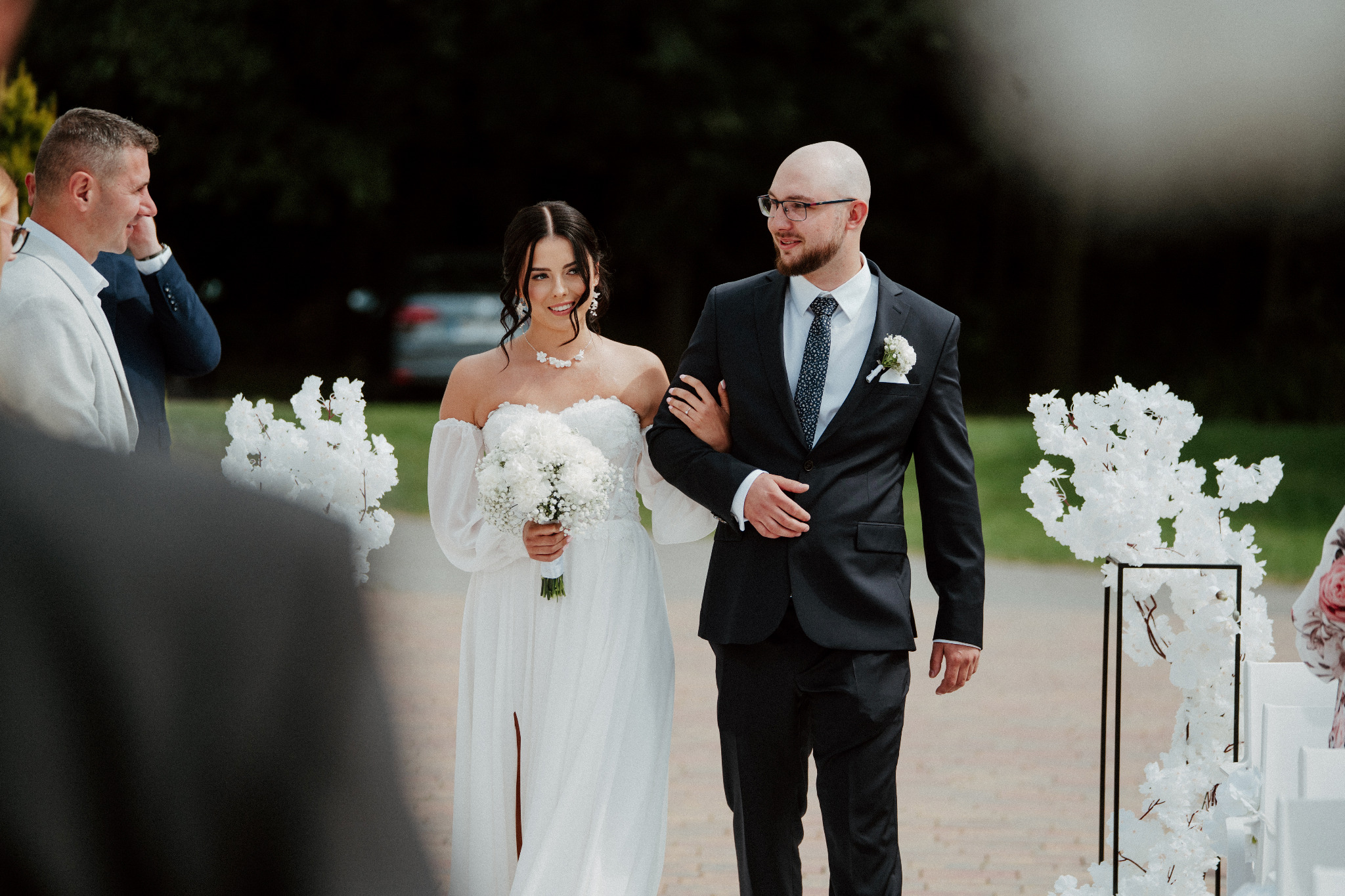 Uśmiechnięta panna młoda w białej sukni i pan młody w garniturze idą alejką udekorowaną białymi kwiatami podczas ceremonii ślubnej w plenerze.