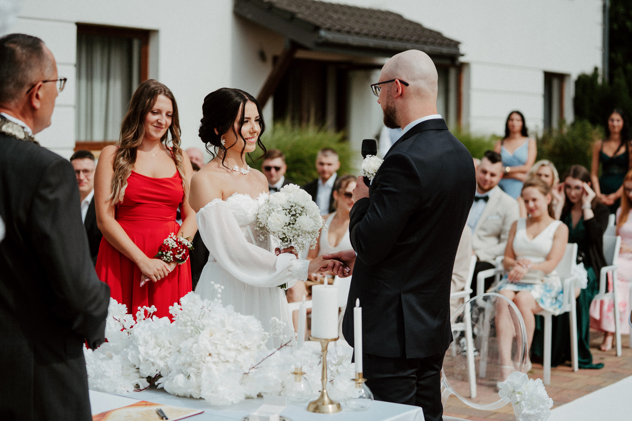 Uśmiechnięta panna młoda w białej sukni trzyma bukiet podczas ceremonii ślubnej na świeżym powietrzu. W tle goście i mężczyzna w garniturze z mikrofonem.