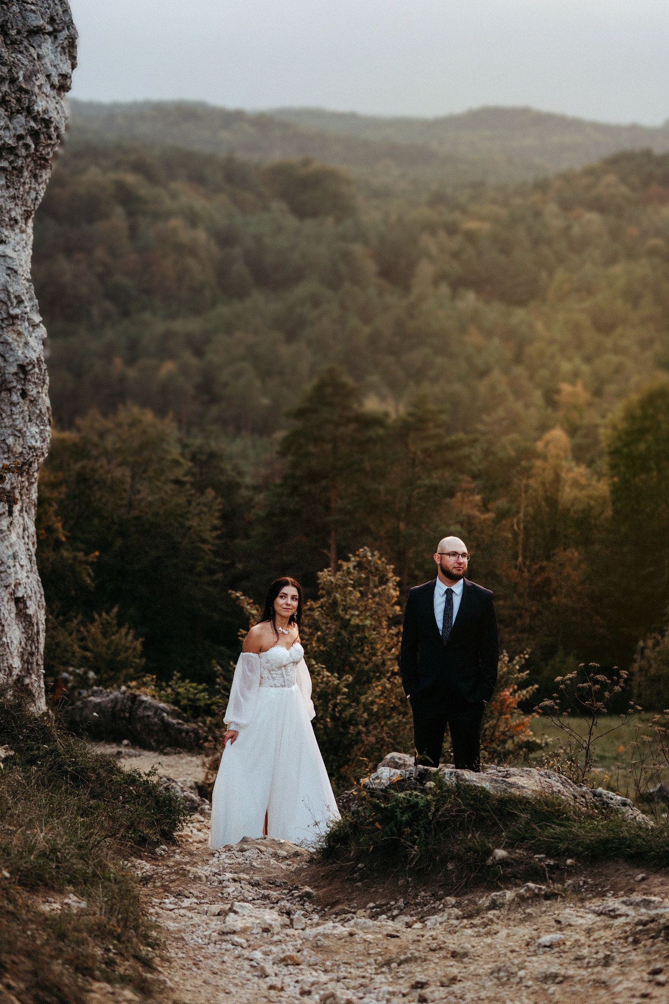 Para młoda w plenerze: panna młoda w białej sukni z odkrytymi ramionami i pan młody w czarnym garniturze na tle leśnego krajobrazu i skał. Romantyczna sceneria.