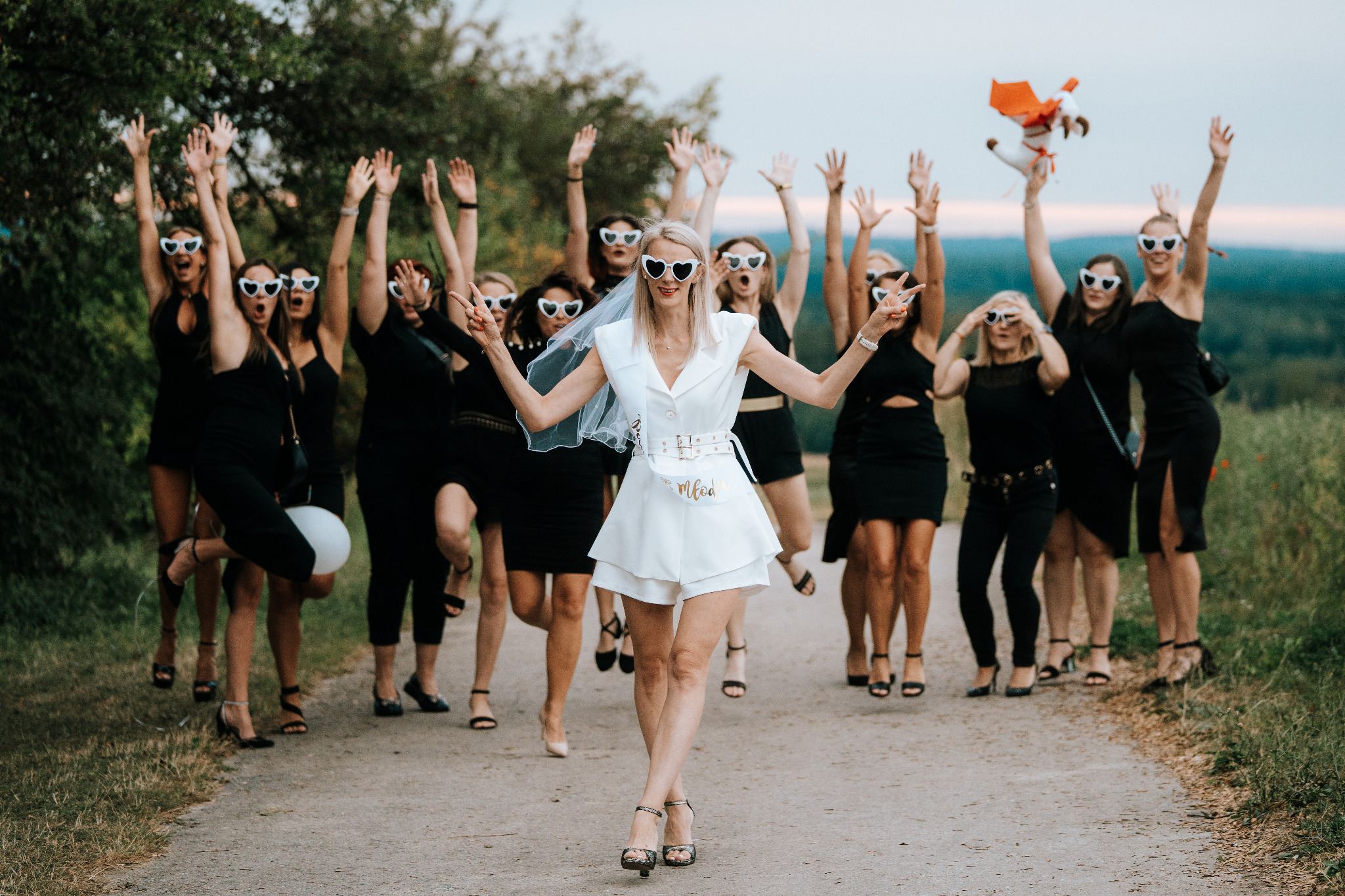 Uśmiechnięta panna młoda w bieli, otoczona druhnami w czerni i okularach w kształcie serc, świętuje wieczór panieński na leśnej drodze.