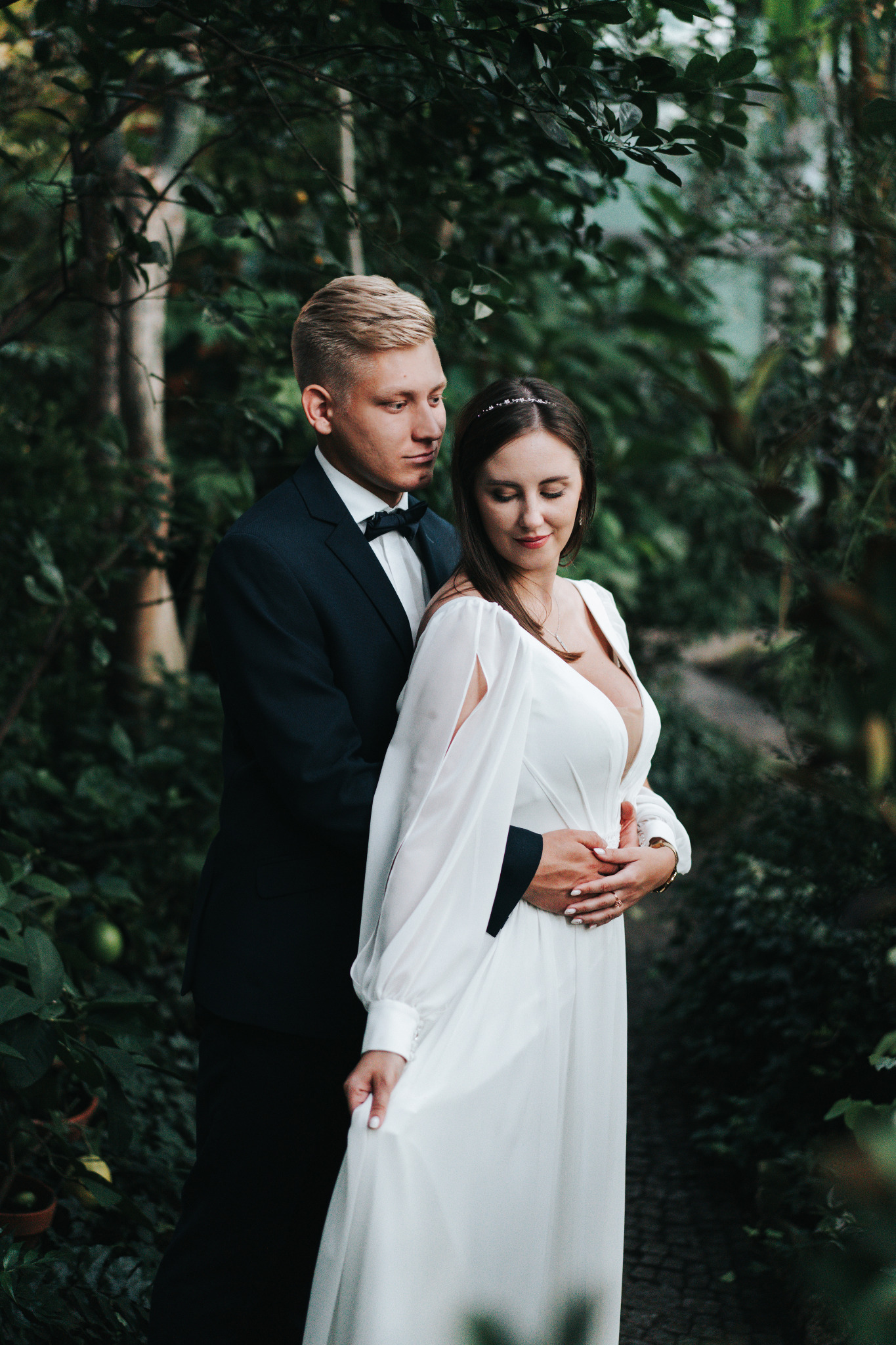 Para młoda w objęciach, on w granatowym smokingu z muchą, ona w białej sukni z długim rękawem, na tle zieleni. Romantyczna sesja w plenerze.