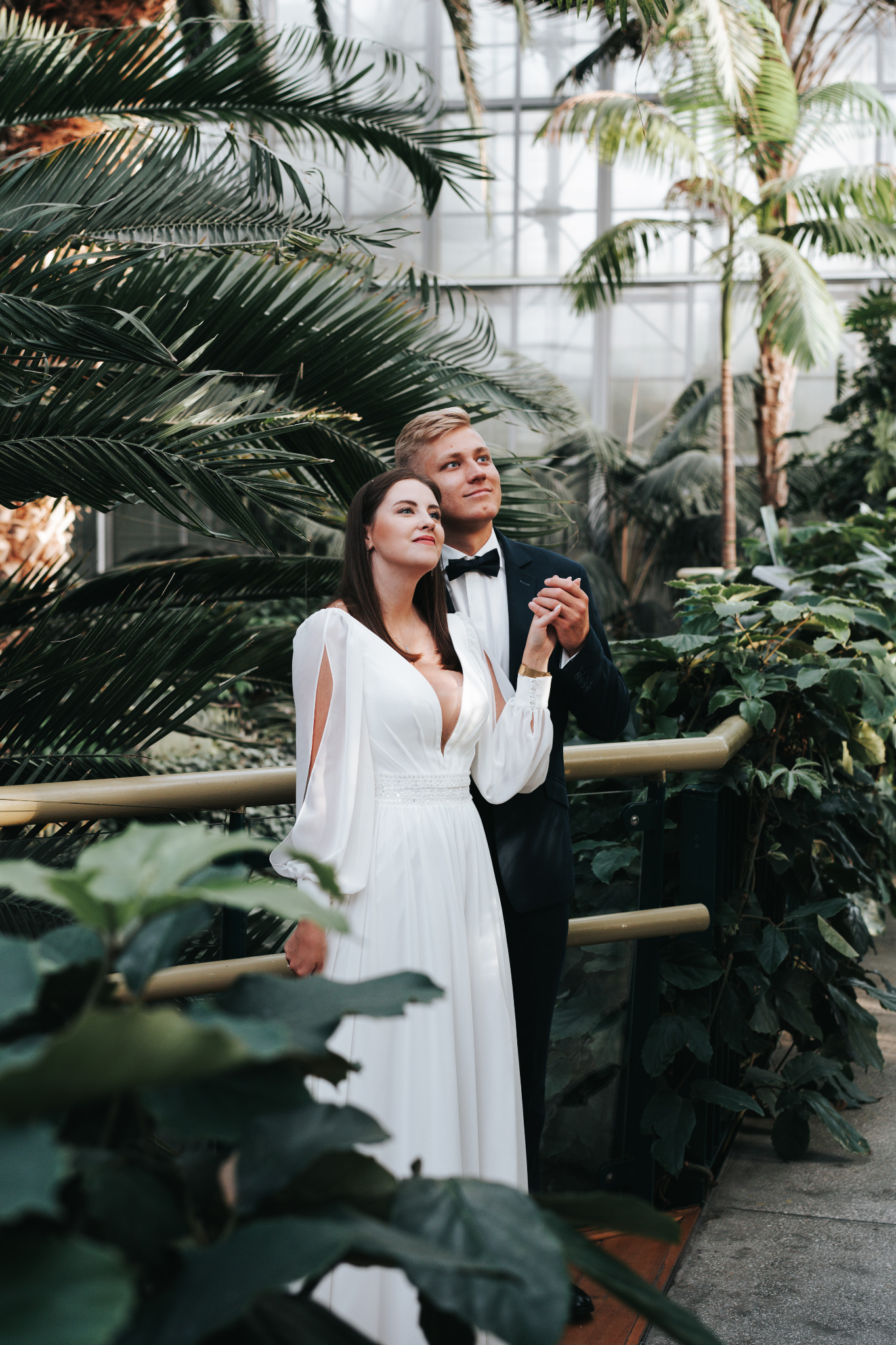 Para w strojach ślubnych, trzymająca się za ręce w tropikalnej scenerii palmiarni w Lędzinach. Panna młoda w białej sukni, pan młody w smokingu.
