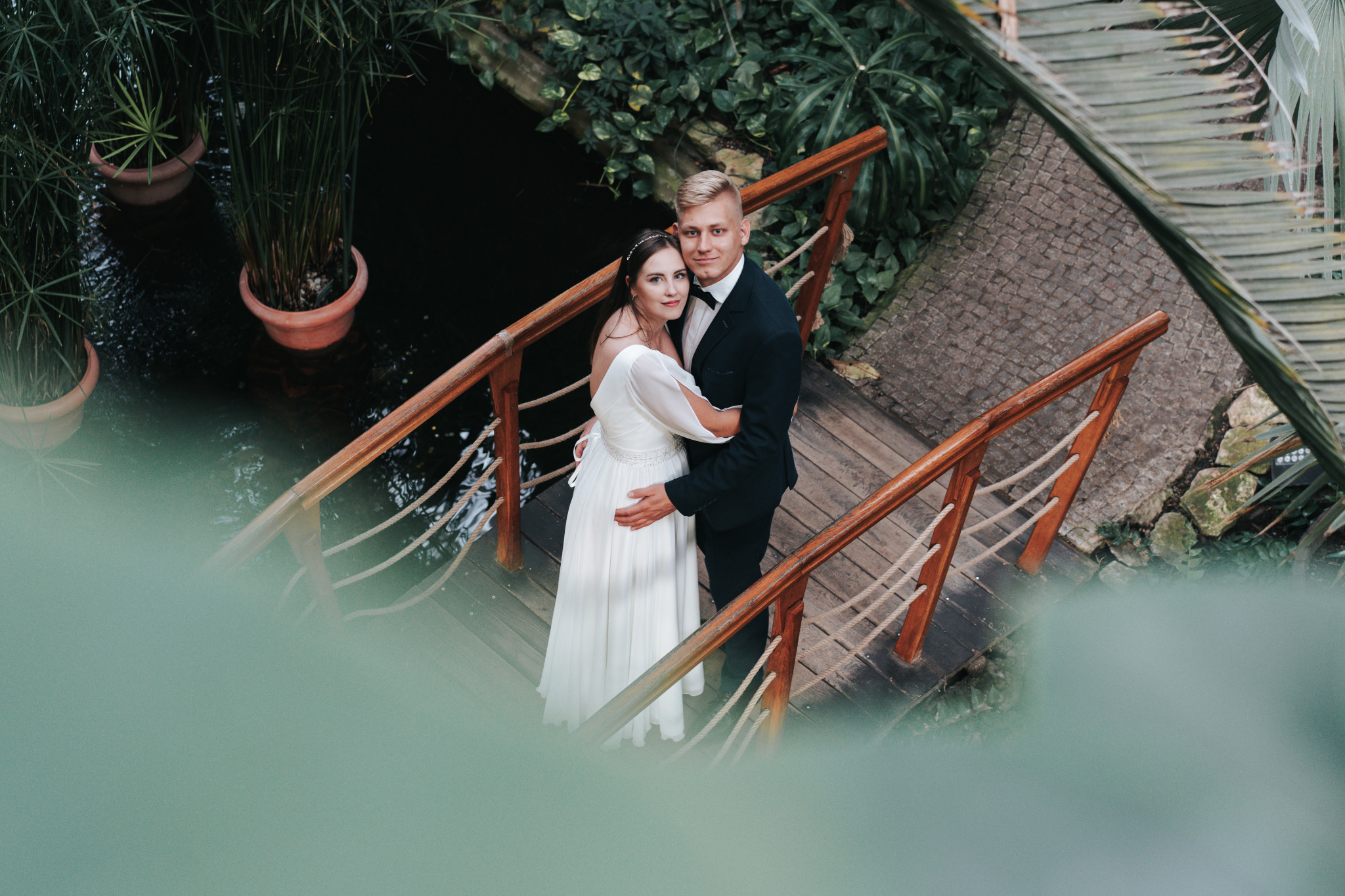 Para młoda na drewnianym mostku w otoczeniu zieleni. Panna młoda w białej sukni, pan młody w czarnym garniturze. Ujęcie z góry, z delikatnym rozmytym pierwszym planem.
