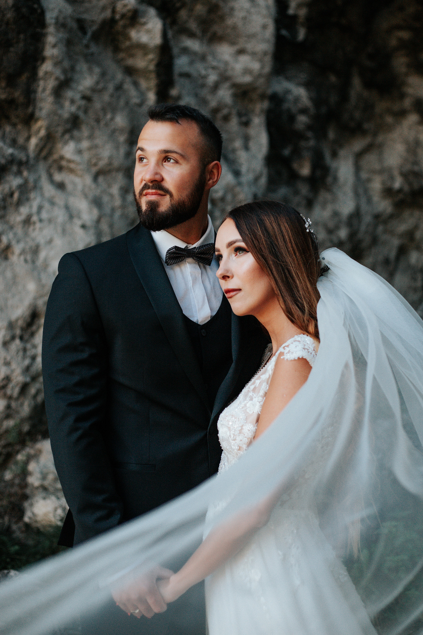 Elegancka para młoda, on w czarnym smokingu z muszką, ona w białej sukni ślubnej z welonem, pozuje na tle skalnej ściany, oboje patrzą w dal.