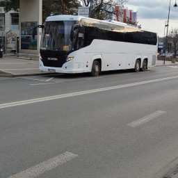 P.H.U. Tomasz Tymrakiewicz - Biały autokar Scania na tle miejskiego krajobrazu z budynkiem z zegarem i flagami. Autobus stoi na ulicy, widoczny z boku, z odbiciami w oknach.