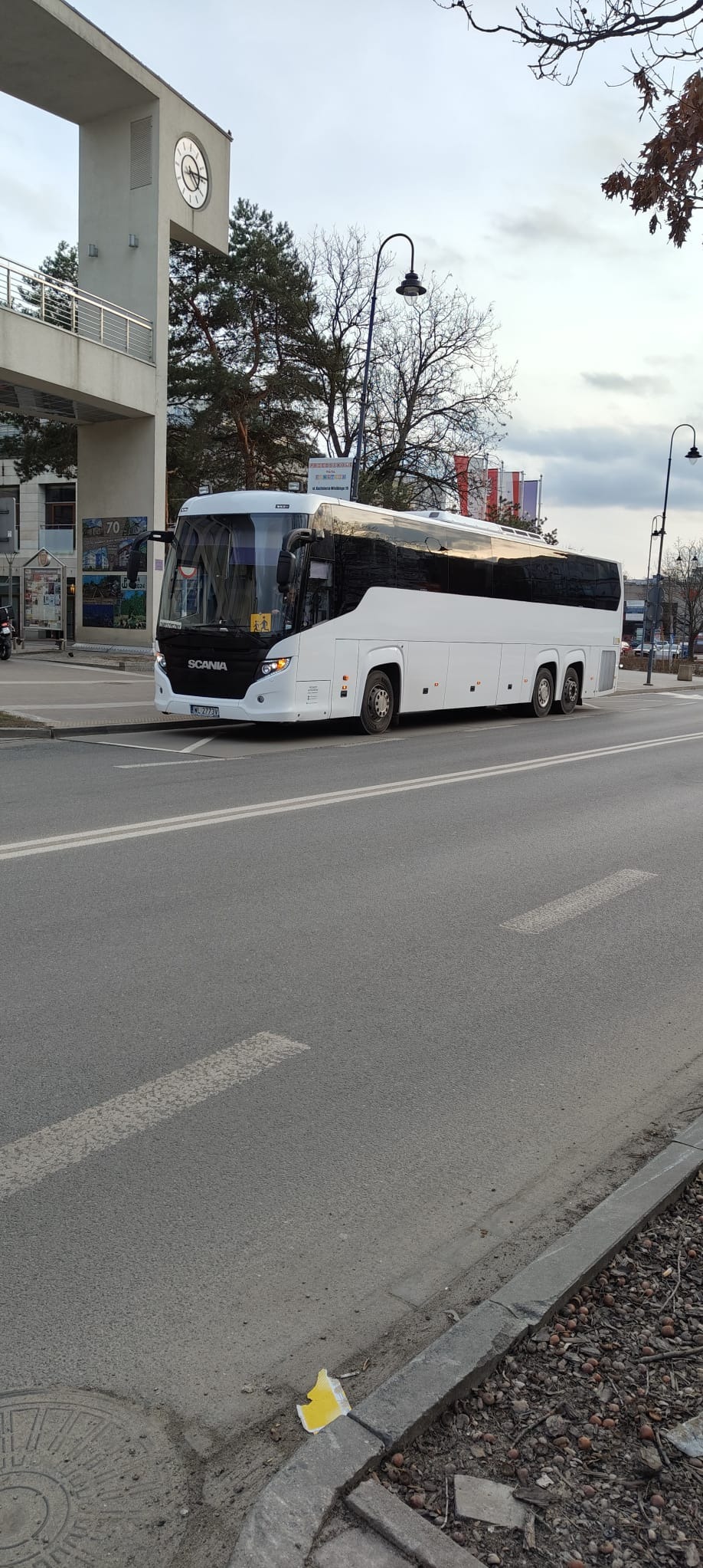Biały autokar Scania na tle miejskiego krajobrazu z budynkiem z zegarem i flagami. Autobus stoi na ulicy, widoczny z boku, z odbiciami w oknach.