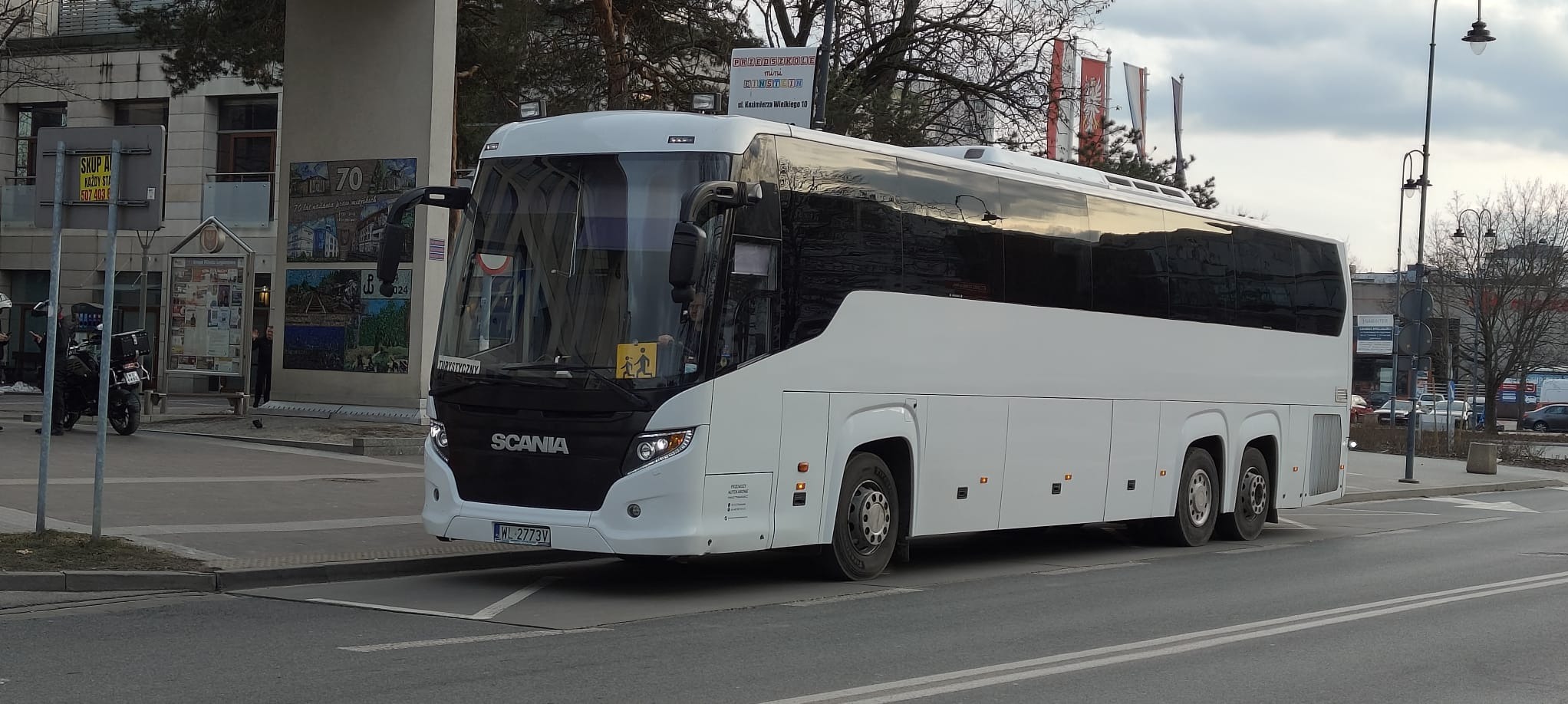 Nowoczesny, biały autokar Scania z tablicą rejestracyjną WL 2773V, zaparkowany na tle miejskiego krajobrazu z elementami zieleni i budynków. Widoczny znak 'Turystyczny'.