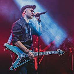 Ginter Media - Wokalista z gitarą typu Flying V na scenie w trakcie koncertu, oświetlony reflektorami w kolorach czerwieni i błękitu. Artysta w czarnym stroju i czapce z daszkiem.