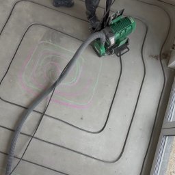 MALGIPS DAWID TRZEŚNIOWSKI - Frezowanie bruzd pod ogrzewanie podłogowe w betonie. Widoczne rury, frezarka i oznaczenia kredą. Praca w toku, z bliska, z góry.