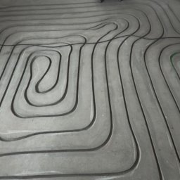 MALGIPS DAWID TRZEŚNIOWSKI - Frezowanie bruzd w posadzce pod ogrzewanie podłogowe. Widoczne równe, wyfrezowane kanały i fragment frezarki. Posadzka przygotowana do montażu rur grzewczych.