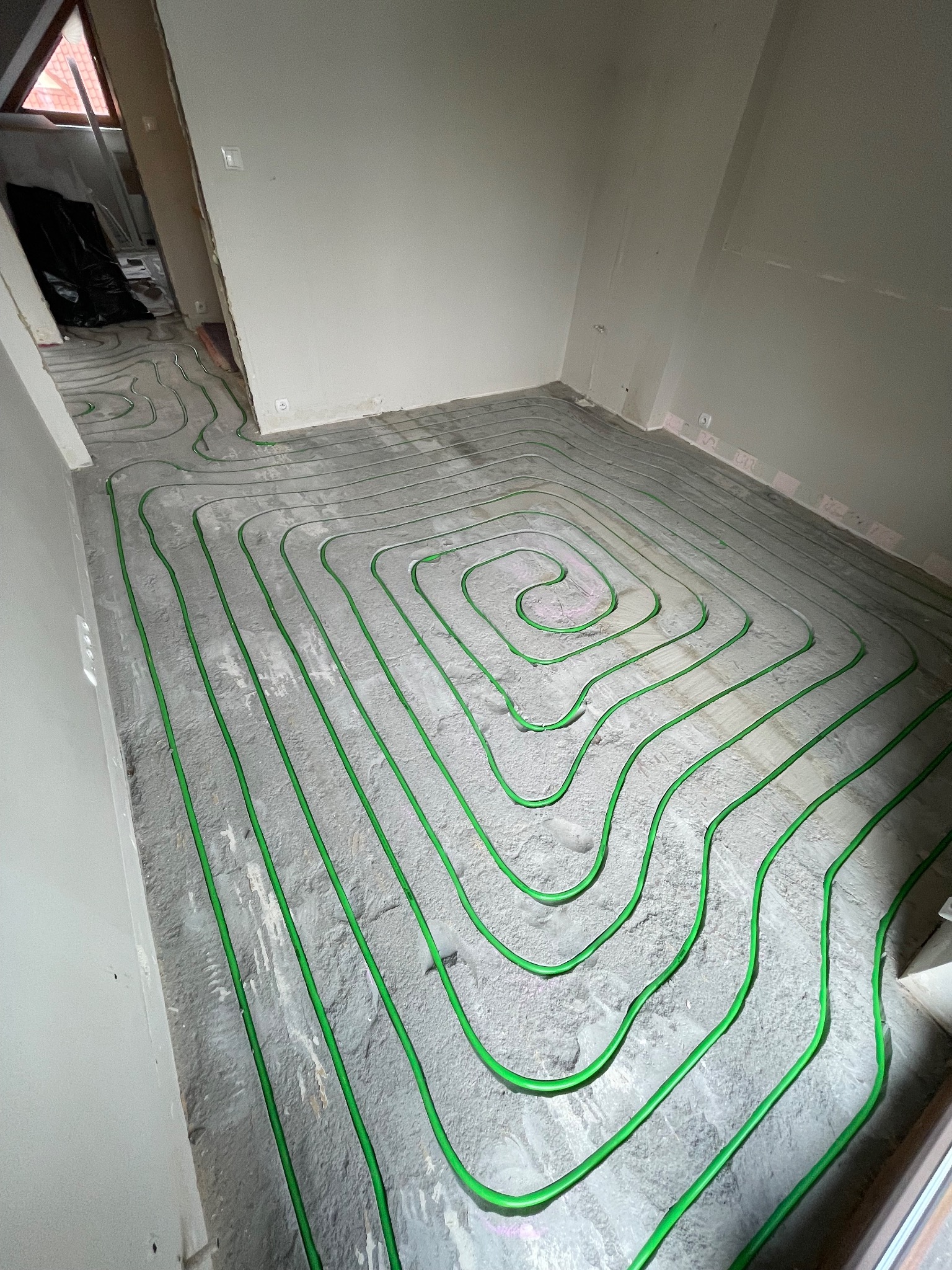 Zielone rurki ogrzewania podłogowego ułożone spiralnie na szarym podłożu w surowym wnętrzu. Widoczne fragmenty ścian i okna.